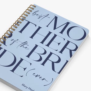 May include: A light blue spiral-bound notebook with dark navy blue text that reads "best MOTHER of the BRIDE (ever)" and the name "Mary Han" printed at the bottom. The notebook features a gold-colored metal wire binding along the left side.