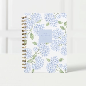 May include: A spiral-bound notebook with a white background and a pattern of blue hydrangea flowers and green leaves. The notebook has a light blue square with the words "OUR WEDDING" and "Save the Date" printed on it.