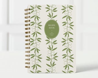 May include: A spiral-bound recipe book with a cream-colored cover featuring a repeating pattern of olive branches in green and white. The center has a green oval with the words "Recipe Book" and "The Anderson Family".