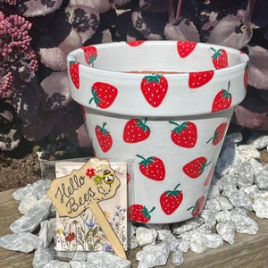 May include: A white ceramic plant pot adorned with red strawberry designs and green leaves. A wooden garden marker with the words "Hello Bees" is in front of the pot. The marker features a bee and flower design. The pot is surrounded by white pebbles.