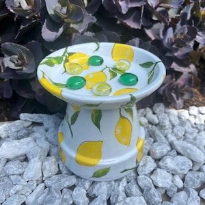 Może przedstawiać: Biała ceramiczna wanienka dla ptaków z ręcznie malowanym wzorem cytryny. Wanienka dla ptaków ma okrągłą górę z płytką misą i podstawą na cokole. W misie znajduje się kilka zielonych i żółtych szklanych kulek. Tło przedstawia fioletowe liście i białe kamienie.