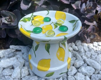 May include: A white ceramic bird bath with a hand-painted lemon design. The bird bath has a round top with a shallow bowl and a pedestal base. The bowl contains several green and yellow glass marbles. The background features purple foliage and white rocks.