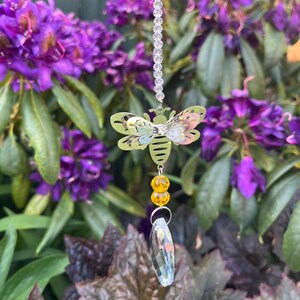 May include: A decorative bee suncatcher with a cut-out design, suspended from a beaded chain. The bee has yellow and pink details, with a cluster of yellow beads and a clear crystal pendant below. The background features purple flowers and green foliage.