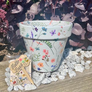 May include: A white ceramic plant pot adorned with a pattern of colourful flowers and dragonflies. A wooden sign reading "Hello Bees" is placed beside the pot. The pot rests on a bed of white stones.