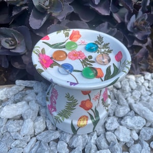 May include: A white, pedestal-style dish with a floral design featuring pink, orange, and red flowers and green leaves. The dish holds several colourful glass marbles. The dish is surrounded by white rocks and purple foliage.