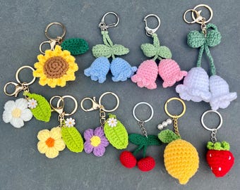 May include: A collection of handmade crochet keychains featuring various floral and fruit designs. The keychains include a sunflower, daisies, tulips, cherries, a lemon, and a strawberry. Each keychain has a metal ring for attaching keys.