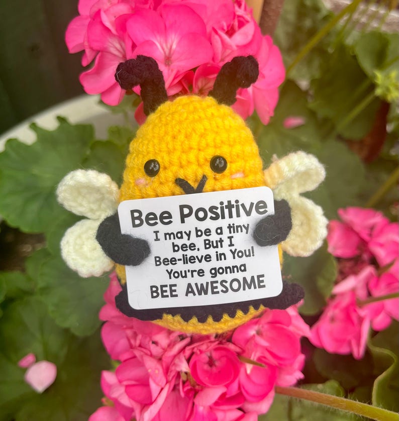 Może przedstawiać: Szydełkowa ż&oacute;łta pszczoła z czarnymi czułkami i skrzydłami, trzymająca znak z napisem "Bee Positive". Na znaku widnieje r&oacute;wnież napis "I may be a tiny bee, But I Bee-lieve in You! You're gonna BEE AWESOME". Pszczoła otoczona jest r&oacute;żowymi kwiatami.