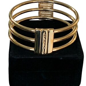 Puede incluir: Un brazalete dorado con tres bandas paralelas. El brazalete presenta detalles rectangulares con texto grabado. La joya se exhibe sobre una superficie negra.