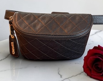 May include: A dark brown quilted belt bag with a textured leather finish and a gold-toned metal charm hanging from the side. The charm features the text CHANEL in gold lettering. The bag has a curved top flap and an adjustable strap attached to the sides.