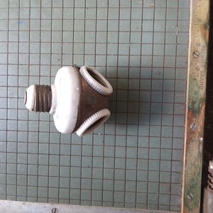 Puede incluir: Un componente eléctrico antiguo con un cuerpo de porcelana blanca y una base de metal marrón. El componente tiene una base de rosca y tres aisladores blancos en forma de anillo. El artículo está colocado sobre una superficie cuadriculada.