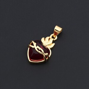 May include: A gold-toned pendant featuring a deep red heart-shaped stone. The heart is encircled by a gold-toned crown of thorns and topped with a stylized gold-toned flame element. A small gold-toned bail is attached to the top for hanging.