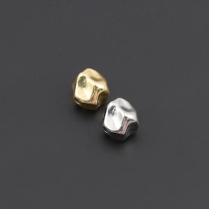May include: Two small, irregular, pebble-shaped metal objects. One object has a polished gold-tone finish, and the other has a polished silver-tone finish. Both items feature a smooth, rounded, and slightly dented surface with reflective highlights.