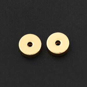 May include: Two small, round, gold-colored metal components with a central hole. The objects are set against a dark background, highlighting their polished surface and simple design. These components could be used in jewelry or other craft projects.
