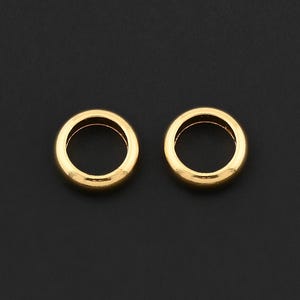 May include: Two small, gold-colored, circular jewelry components with a smooth, polished finish. The components are set against a dark background, highlighting their shape and color. These could be earring findings or other jewelry parts.