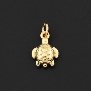 May include: A small, gold-colored turtle charm with a textured shell and flippers. The charm has a loop at the top for attaching to a necklace or bracelet. The turtle is centered on a black background.