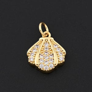Puede incluir: Un colgante dorado con forma de concha, con un lazo para una cadena. El colgante está incrustado con pequeños cristales transparentes, que añaden brillo. El diseño es detallado y elegante, para joyería.