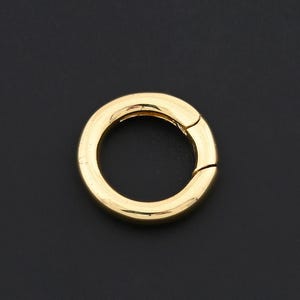 May include: A gold-colored, circular clasp with a hinged opening. The clasp is set against a dark background, highlighting its polished finish and simple design. This jewelry finding is suitable for various crafting projects.