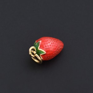 Puede incluir: Un charm de fresa roja con hojas verdes y un lazo dorado. El charm tiene una superficie texturizada y está sobre un fondo oscuro. Esta pieza de joyería es un accesorio con temática de frutas.