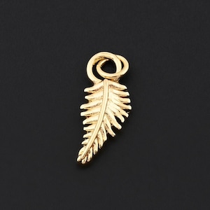 May include: A small, gold-colored pendant in the shape of a fern leaf. The pendant has intricate detailing, with each leaf segment clearly defined. The bail is a simple loop. The background is a solid black.