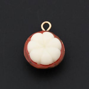 May include: A charm shaped like a mangosteen fruit, featuring a white, flower-like center surrounded by a reddish-brown rind. The charm has a gold-colored loop at the top for attaching to jewelry. The background is a solid black.