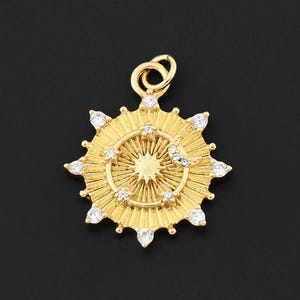 May include: A gold pendant with a sunburst design. The pendant features a central starburst pattern surrounded by a ring of small, clear gemstones. The pendant has a loop at the top for a chain.