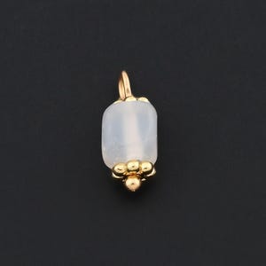May include: A gold-colored pendant with a rectangular, light-colored stone. The pendant has a small loop at the top and a cluster of small gold beads at the bottom. The background is a solid black.