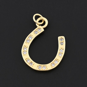 May include: A gold-colored horseshoe pendant with small, clear gemstones set along its curve. The pendant has a loop at the top for attaching to a chain or bracelet. The background is a solid black.
