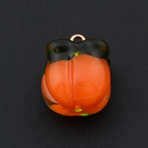Puede incluir: Un pequeño dije con forma de calabaza naranja con una parte superior verde oscuro y un lazo dorado. El dije tiene un acabado brillante y probablemente está hecho de vidrio o resina. El fondo es negro sólido.