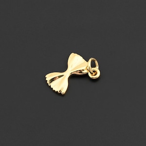 Pode incluir: Um pingente dourado em forma de gravata borboleta. O pingente tem um pequeno laço na parte superior para prender a uma corrente. O design da gravata borboleta tem bordas detalhadas e um acabamento polido. O fundo é cinza escuro sólido.