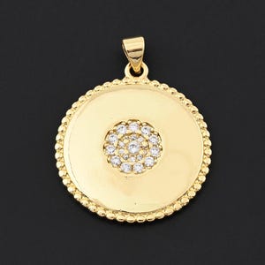 Puede incluir: Colgante dorado con diseño circular. El colgante presenta un grupo central de pequeñas piedras transparentes rodeadas por un borde de cuentas. El colgante tiene un lazo en la parte superior para una cadena.