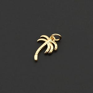 Op de afbeelding: Een kleine, goudkleurige palmboombedel. De bedel heeft een gedetailleerd ontwerp van een palmboom met een getextureerde stam en bladeren. De bedel heeft een kleine lus aan de bovenkant om aan een ketting of armband te bevestigen. De bedel staat tegen een donkere achtergrond.