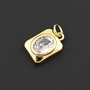 May include: A gold-colored pendant with a rectangular frame and an oval, clear gemstone in the center. The pendant has a small loop at the top for attaching to a chain. The background is a solid black.