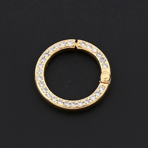 May include: A gold-colored, circular clasp with a hinged opening. The clasp is encrusted with small, clear gemstones around the outer edge. The background is a solid black.