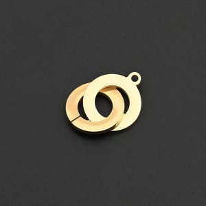以下が含まれることがあります： 2つの円が組み合わさったゴールドカラーのペンダント。1つは小さな開口部があります。ペンダントの上部には、チェーンや他のジュエリーコンポーネントに取り付けるための小さなループが付いています。背景は無地の黒です。