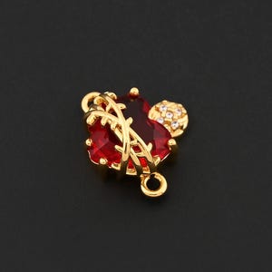 May include: A gold-colored charm with a red gemstone heart shape, accented with a smaller, round, clear-stone cluster. The charm has a loop at the top and bottom for attachment. The design is set against a black background.