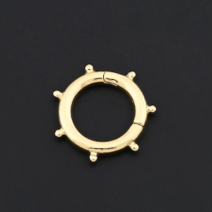May include: A gold-colored, circular clasp with a hinged opening. The clasp features small, evenly spaced decorative studs around its outer edge. The background is a solid black.