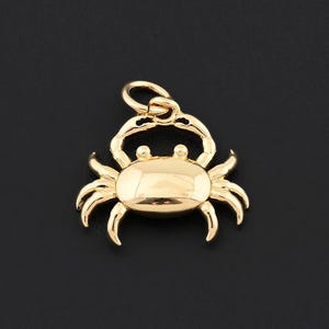 May include: A gold-colored crab charm with a loop for attaching to a necklace or bracelet. The crab has a smooth, polished surface and detailed claws and eyes. The charm is set against a black background.