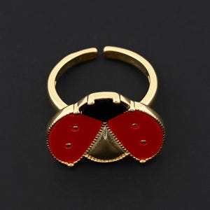 May include: A gold-colored adjustable ring featuring a ladybug design. The ladybug has a red enamel finish with black spots and a gold-colored body. The ring is set against a black background.