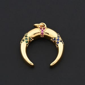 K&ouml;nnte beinhalten: Goldfarbener Halbmond-Anh&auml;nger mit bunten Edelsteinen. Der Anh&auml;nger hat eine gebogene Form mit einer Schlaufe f&uuml;r eine Kette. Die Edelsteine sind in Rot, Gr&uuml;n und Blau gehalten und verleihen dem Gold Farbe. Der Anh&auml;nger ist vor dunklem Hintergrund platziert.