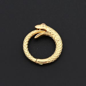May include: A gold-coloured snake-shaped clasp with a textured surface. The snake's head is at the top, with a small gem on its head. The clasp is circular and has a hinged opening.