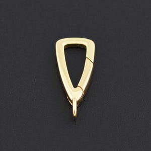 May include: A gold-colored, triangular clasp with a spring-loaded closure. The clasp has a small loop at the bottom for attaching to other jewelry components. The background is a solid black.