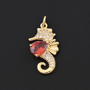 May include: A gold-colored seahorse pendant with a red teardrop-shaped gemstone and clear crystals. The seahorse has a curved tail and a detailed head, with the gemstone positioned near the center. The pendant is designed for jewelry.