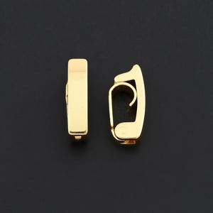 Könnte beinhalten: Zwei goldfarbene Ohrringe auf schwarzem Hintergrund. Ein Ohrring hat eine rechteckige Stabform, der andere ein gebogenes Design mit einem kleinen Haken. Die Ohrringe bestehen aus glänzendem, reflektierendem Metall.