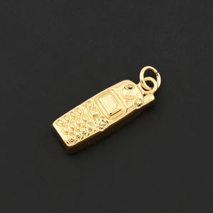 May include: A gold-colored pendant in the shape of a vintage flip phone. The pendant has detailed textures on the front and a small loop at the top for attaching to a chain or bracelet. The background is a solid black.