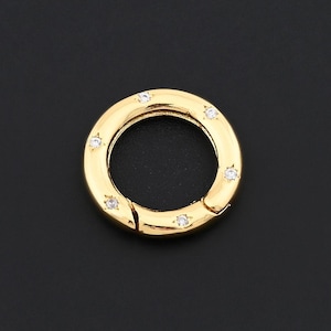 May include: A gold-colored circular clasp with small, clear gemstones evenly spaced around its outer edge. The clasp is open, revealing a hinged mechanism. The background is a solid black.