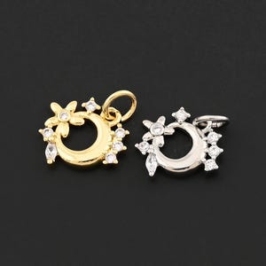May include: Two pendants, one gold-toned and one silver-toned, each featuring a crescent moon design adorned with small, clear gemstones and a flower. The pendants are displayed against a dark background.