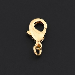 May include: A single gold-colored lobster clasp, a jewelry component, is displayed against a dark background. The clasp is in a closed position, showing its curved design and spring-loaded mechanism. The metal has a shiny, reflective surface.