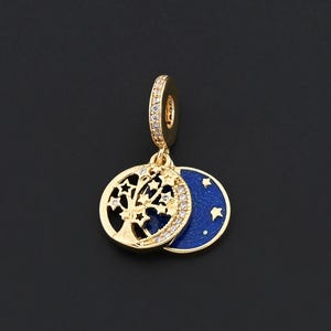 May include: A gold-coloured charm featuring a tree of life design and a blue enamel moon with stars. The charm has a small loop with clear stones. The tree of life charm is round and has a cut-out design.