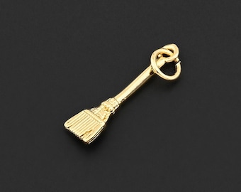Puede incluir: Un dije dorado con forma de escoba. La escoba tiene un mango y cerdas detalladas, con un pequeño lazo en la parte superior para sujetarlo a un collar o pulsera. El dije está sobre un fondo oscuro.