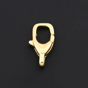 May include: A gold-colored lobster clasp, a jewelry component, is displayed against a black background. The clasp is a shiny, metallic piece with a spring-loaded mechanism for secure closure. It is designed for attaching to necklaces, bracelets, or other jewelry.